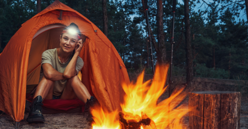 Žena sedí ve stanu s čelovkou vedle ohně v lese