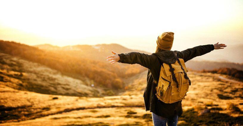 Osoba s batohem stojí na louce s roztaženýma rukama